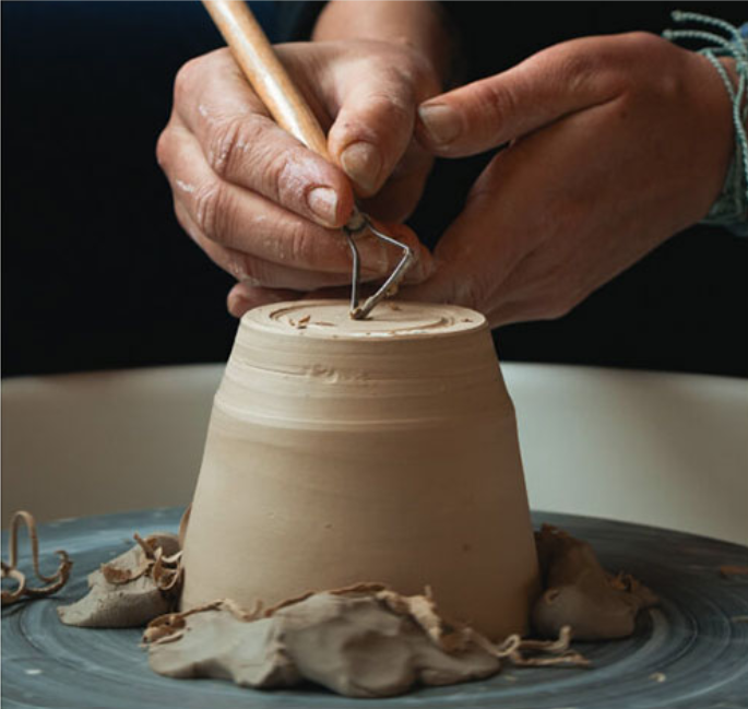 Hands sculpting claypot using a pottery sculpturing instrument.